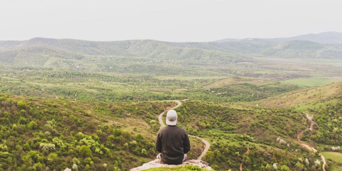 A person sits on a hill overlooking a vast, green valley under a clear sky.
