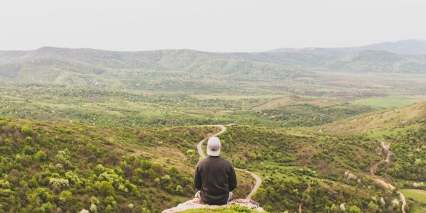 A person sits on a hill overlooking a vast, green valley under a clear sky.